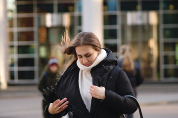 Fototapeta premium Portrait of a confident busy modern pretty woman. Girl hurries and fastens outerwear on the go. Hair develops in the wind. Background business center