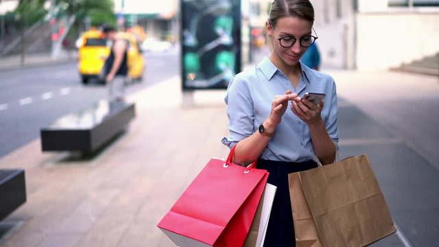 Stylish young woman with bags in hands doing shopping and talking on smartphone device while standing on street of city.Hipster girl calling on phone and checking time on smartwatch to appoint meeting