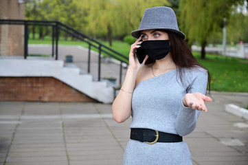Photo of a cute young Caucasian brunette girl in a hat talking on the phone in a black medical mask. Model posing in a city park quarantined outdoors.