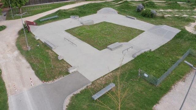 Aerial shot of an empty skatepark. Drone flies over the concrete park with green grass in the middle. Park is closed due to corona lockdown.