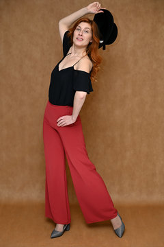 Photo full-length portrait on a beige background Caucasian pretty young woman in a black blouse, red trousers. Model posing with a hat in her hands with long beautiful red hair.