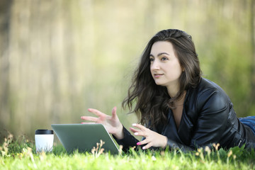 Young woman in a black jacked with coffee cup and laptop working outside in a park. Remote work. Digital work. Distance learning