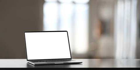 Photo of white blank screen computer laptop putting on working desk over comfortable living room as...