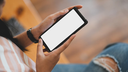 Cropped image of beautiful woman holding a cropped smartphone with white blank screen while sitting in the comfortable sitting room.