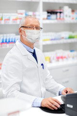 medicine, healthcare and people concept - senior apothecary wearing face protective medical mask for protection from virus disease at pharmacy cash register