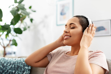 people, technology and leisure concept - happy young african american woman with headphones sitting on sofa and listening to music at home