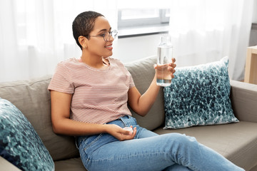 people, sustainability and leisure concept - happy smiling african american young woman sitting on sofa and drinking water from glass bottle at home