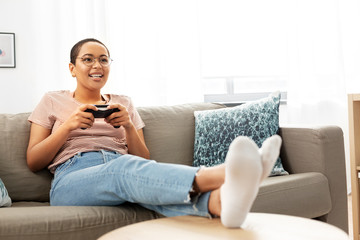 Obraz premium people and leisure concept - happy smiling african american young woman with gamepad playing video game at home