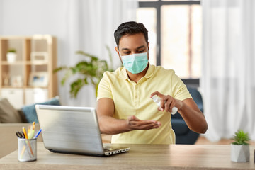 quarantine, remote job and pandemic concept - happy indian man wearing face protective medical mask for protection from virus disease using hand sanitizer with laptop computer working at home office
