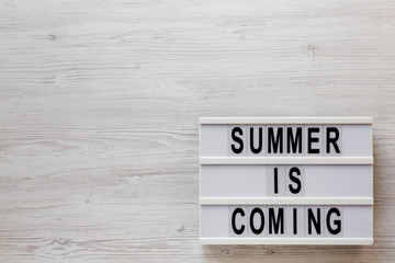 'Summer is coming' text on a lightbox on a white wooden background, top view. Flat lay, overhead, from above. Copy space.