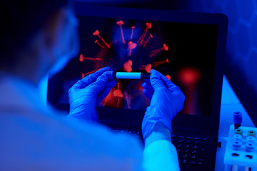 medicine, healthcare and virus concept - close up of hand in protective medical glove holding beaker with blood test over coronavirus on laptop computer screen at laboratory