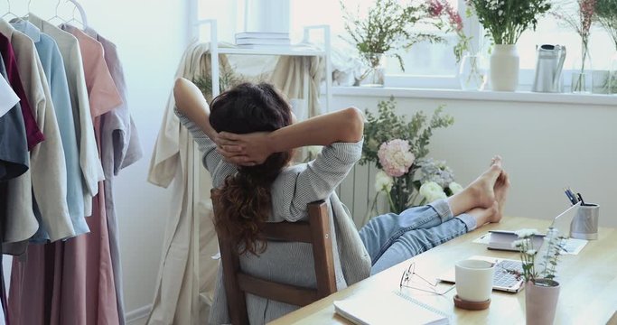 Back rear view of female fashion designer relaxing at workplace putting hands behind head with feet on table. Young business woman chilling sitting on chair taking break at work in feminine studio.