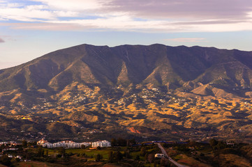 Fototapeta premium A panorama of a mountain range illuminated by the contrasting light of the sun with villages of white low-rise buildings, roads and a golf course in the foreground