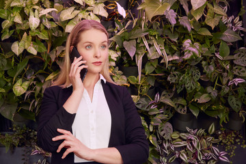 Beautiful blonde businesswoman standing in front of the plant. She is on the phone.