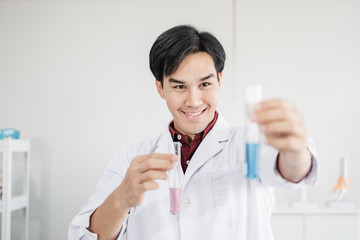 A young male scientist with black hair smiling wearing a white lab coat smiling and holding up test tubes with blue solution in a laboratory setting.