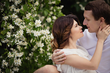 Fototapeta premium Young man and woman couple in a blooming garden near jasmine bush. Tender holding each other. Spring lovestory. Brown-haired girl with long hairs with her boyfriend or husband. Young couple on a date