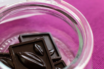 Pieces of handmade chocolate are in a glass jar on a lilac background, close-up, selective focus.