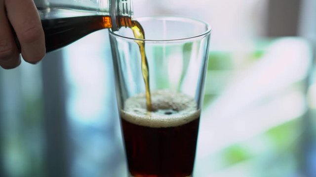 Glass of pop cola being poured into glass on reflective table.