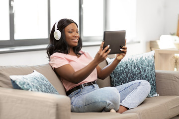 Obraz premium people, technology and leisure concept - happy young african american woman with tablet pc computer and headphones sitting on sofa and listening to music at home