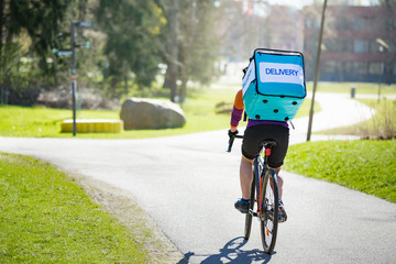 Naklejka premium Woman in helmet and protective mask with big food delivery backpack riding bike on the street. Sunny spring day in the city. Portrait of a female courier in a medical mask.