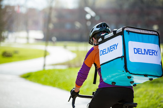 Woman In Helmet And Protective Mask With Big Food Delivery Backpack Riding Bike On The Street. Sunny Spring Day In The City. Portrait Of A Female Courier In A Medical Mask.