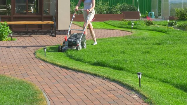 A woman mows green juicy grass with a lawnmower in the backyard of the house. Beautiful lawn and winding garden path. Front view