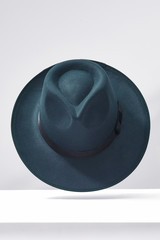 Subject shot of a sea-green fedora hat decorated with a black silk band with a bow. The stylish hat is fixed in the air under the white surface against the gray backdrop.