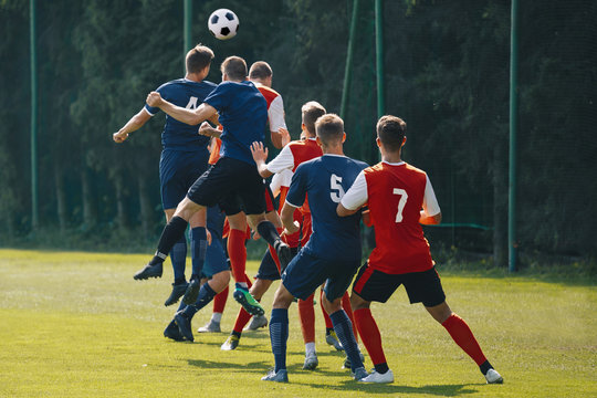 Soccer Players Heading The Ball In Competition. Football Adult Game. Players In Two Teams Compete For The Ball. Footballers Jumping High On The Grass Pitch