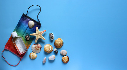 Summer vacation fat lay with protective mask painted in rainbow colors, antiseptic, seashells, pebbles and starfish on blue background. Travel and coronavirus covid-19 concept. Banner