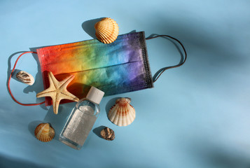 Summer vacation fat lay with protective mask painted in rainbow colors, antiseptic, seashells, pebbles and starfish on blue background backlit by the sun. Travel and coronavirus covid-19. Copy space