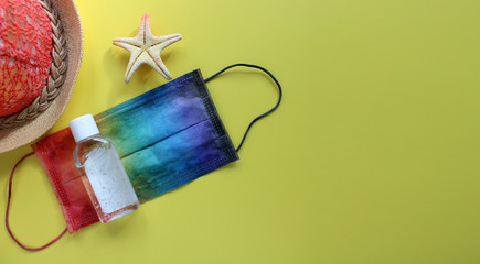 Summer vacation flat lay with protective mask painted in rainbow colors, antiseptic, beach hat and starfish on yellow background. Travel and coronavirus covid-19. Copy space