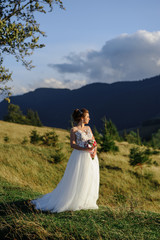 Obraz na płótnie Canvas The bride herself with a wedding bouquet poses against the backdrop of the mountains at sunset.