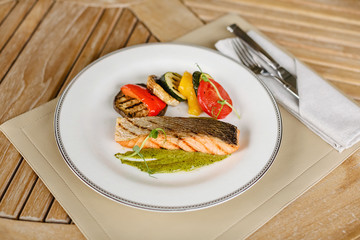 close-up of a round white plate on a wooden table. Fish steak grilled salmon and vegetables eggplant, pepper, tomato grilled in the restaurant