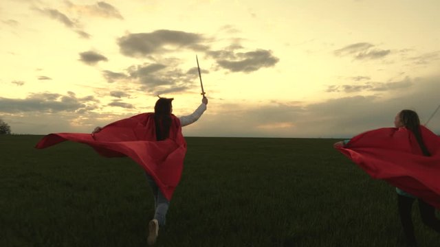 free young girls play super heroes. girls in red cloaks run with swords in hand across field playing medieval knights. children fight with toy sword. healthy children play knights. happy childhood