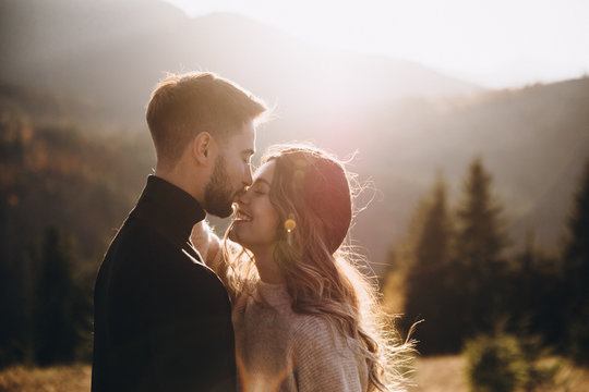 Stylish model couple in the autumn mountains. A young guy and a girl hug and kiss on a background of a forest and mountain peaks at sunset.