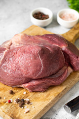 Beef ham on a wooden Board on the light gray kitchen table. Raw beef meat. A large chunk of meat lies on a chopping Board, next to bowls of salt, pepper and vegetable oil