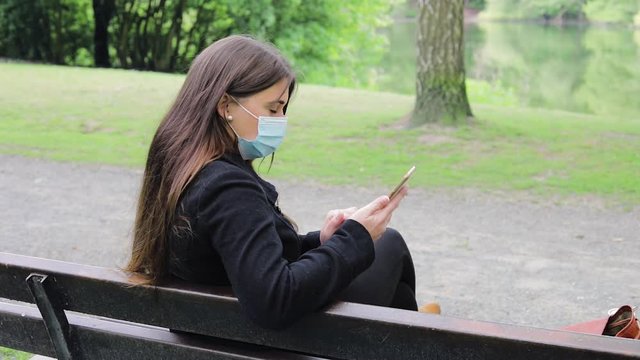 young woman with Corona mask on her phone in a park on a bench