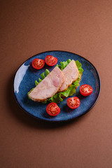 Sandwich with turkey ham meat, green salad and fresh cherry tomatoes slices on blue shiny plate, brown minimal background, angle view