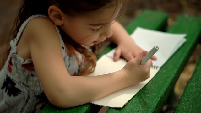 Little Girl Draws On Paper In Park.Cute Small Kid Girl Enjoying Creative Art.Adorable Preschool Child Learning Drawing Picture.Daughter Child Painting With Pencil On Creative Book Education Homework.