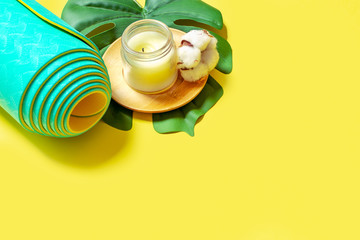 Natural wooden colored wax candle with cotton, green leaf and rolled turquoise mint yoga, pilates mat on the yellow background. Flat lay and space for text. Relax and meditation concept.