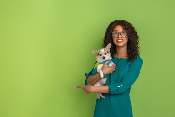 Pointing and smiling cheerful. African-american young woman's portrait isolated on green studio background. Beautiful model with little doggy. Concept of human emotions, facial expression, sales, ad.