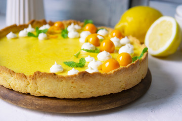 Lemon tart decorated with curd, cream cheese balls, physalis berries, mint leaves. Freshly baked dessert on white textured table with green leaves, lemons. Beautiful citrus pie. Copy space.