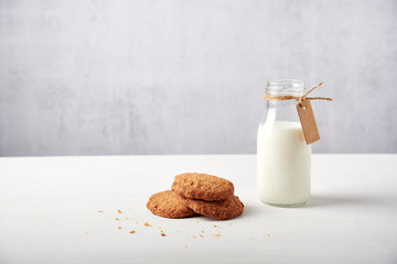 Cookies next to milk bottle with tag on white wooden table against light grey grunge background, a lot of negative space, side view