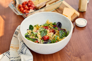 High angle view of bowls with pasta salad and salt near napkin and cutting board with parmesan and baking dish on wooden background