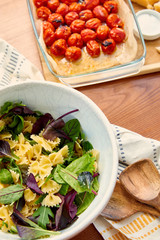 High angle view of bowl with pasta salad ingredients near spatulas, napkin and cutting board with tomatoes in baking dish on wooden background