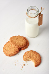 Cookies next to milk bottle with tag on white wooden table, one cookie bitten off. 45 degrees vertical view