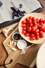 High angle view of plate with tomatoes on cutting board near oven tray with ingredients on wooden background