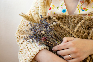 A young woman in a beige sweater holds a knitted mesh bag and a bouquet of dry lavender.