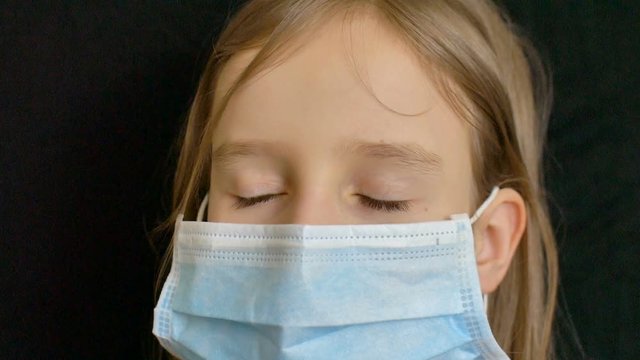 Little blond girl with blue eyes and long hair is posing on black backgound in surgical disposable mask for protection of Coronavirus Covid-19 during self-isolation while staying at home