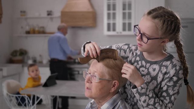 Close-up of caring teenage granddaughter gently combing hair of paralyzed grandmother. Loving grandfather cooking by stove and little grandson watching video for kids on tablet blurred on background